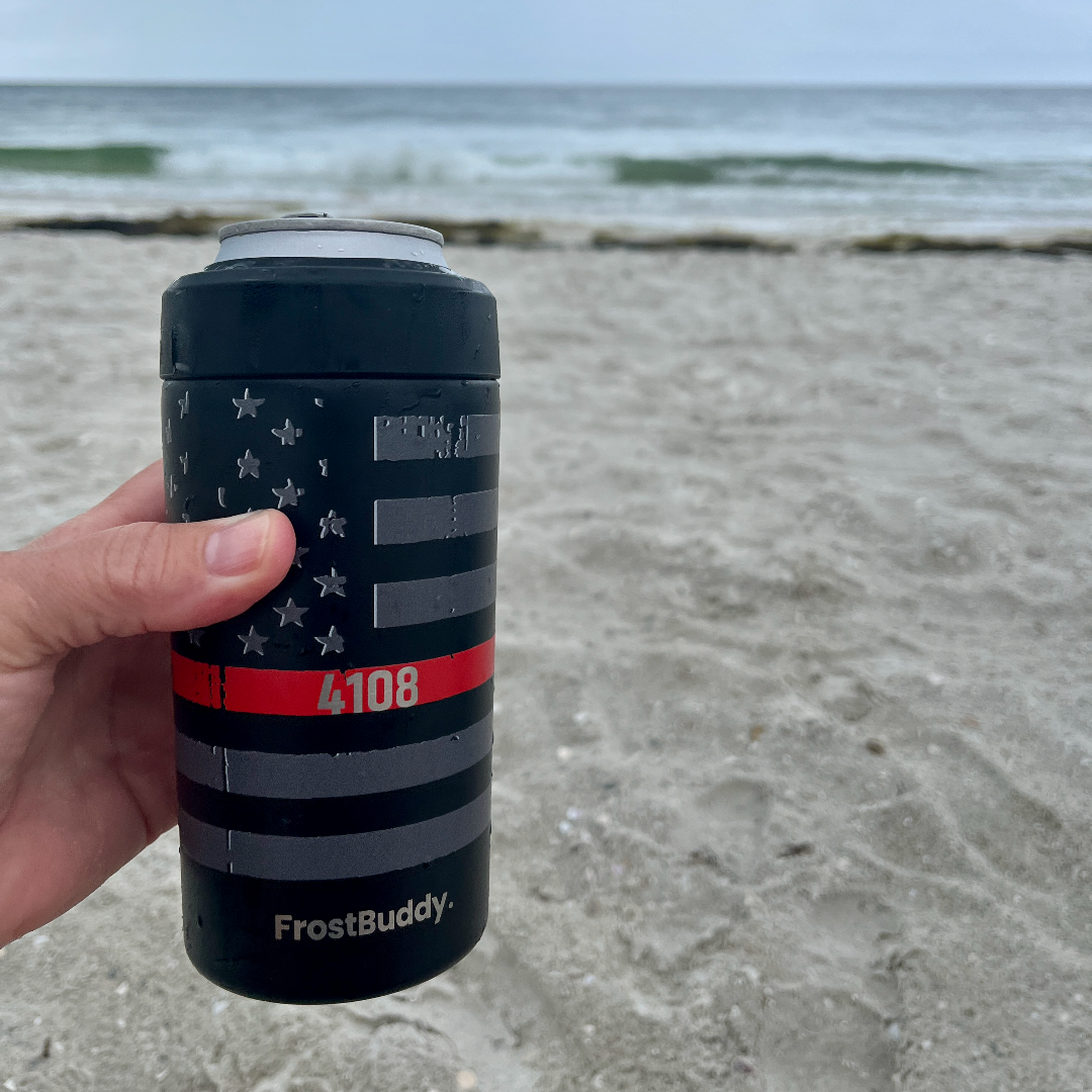 FrostBuddy Universal Can Cooler with firefighter badge number engraving. Custom engraved Firefighter FrostBuddy Universal can cooler with badge number, held at the beach with ocean background — personalized first responder drink holder.